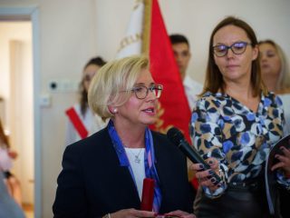 Kobieta w okularach przemawia do mikrofonu obok prowadzącej uroczystość.