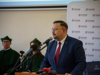 Mężczyzna w granatowym garniturze i czerwonym krawacie przemawia przy mównicy.