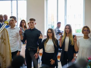 Grupa studentów składa ślubowanie z uniesionymi prawymi dłońmi obok sztandaru uczelni.