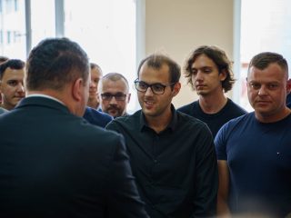 Mężczyzna w garniturze stoi przed grupą uczestników uroczystości, wśród których jest mężczyzna w okularach i czarnej koszuli.