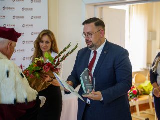 Mężczyzna z czerwoną krawatką trzyma szklaną statuetkę i dokument podczas spotkania z rektorem.