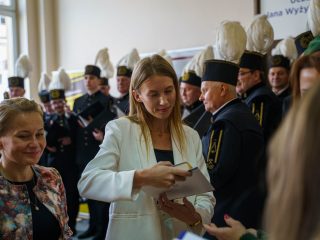 Kobieta w białej marynarce trzyma książkę i dokument podczas wręczania wyróżnień.