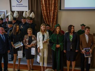 Grupa wyróżnionych osób pozuje z dokumentami i książkami obok przedstawicieli uczelni.