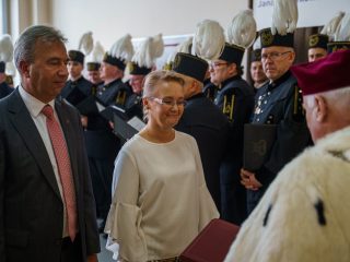 Kobieta w białej bluzce i mężczyzna w garniturze stoją przed rektorem podczas uroczystości.