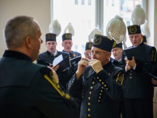 Członek chóru w mundurze górniczym gra na harmonijce ustnej przed pozostałymi chórzystami.