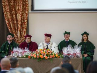 Przedstawiciele uczelni w togach siedzą za stołem z kwiatami i upominkami.