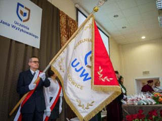 Mężczyzna z biało-czerwoną szarfą trzyma sztandar UJW obok stołu prezydialnego.