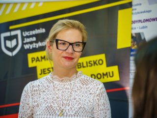 Kobieta w okularach i białej koronkowej bluzce pozuje na tle banera UJW.