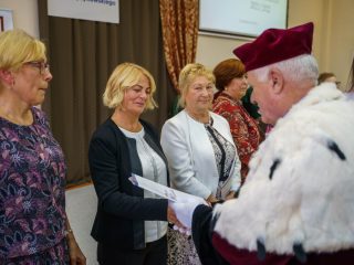 Rektor wręcza dokument kobiecie w ciemnej marynarce stojącej wśród uczestniczek.