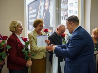 Mężczyzna w niebieskim garniturze gratuluje kobiecie w jasnej marynarce trzymającej dyplom.