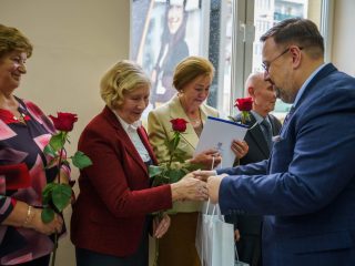 Kobieta w bordowym żakiecie z czerwoną różą ściska dłoń mężczyzny wręczającego upominki.