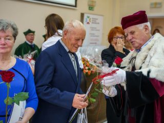 Rektor wręcza czerwoną różę uśmiechniętemu mężczyźnie w garniturze.