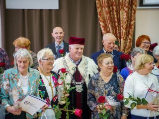 Rektor pozuje wśród uśmiechniętych uczestników z różami i upominkami.