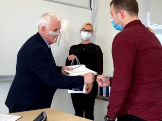 Mężczyzna w bordowej koszuli odbiera gratulacje, dotykając pięścią dłoni wręczającego nagrody.
