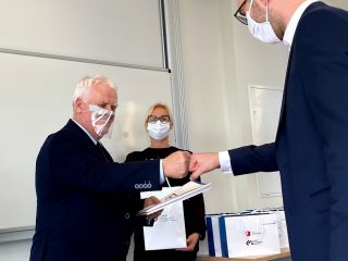 Mężczyzna w granatowym garniturze i okularach wita się dotknięciem pięści z organizatorem uroczystości.