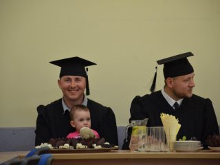 Dwaj absolwenci w togach i biretach siedzą przy stole; jeden trzyma małe dziecko.