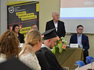Siwowłosy mężczyzna przemawia przed absolwentami, stojąc za stołem z kwiatami.