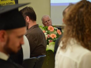 Widok między uczestnikami na siwowłosego mężczyznę siedzącego za bukietem kwiatów.