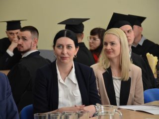 Dwie kobiety siedzą przy stole, a za nimi absolwenci w togach i biretach.