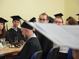 Absolwenci w biretach siedzą przy stole z poczęstunkiem i słuchają wystąpienia.