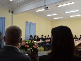 Szeroki widok sali z absolwentami i gośćmi siedzącymi przy długich stołach.