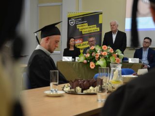 Absolwent w todze siedzi przy stole, a w tle siwowłosy mężczyzna przemawia za kompozycją kwiatową.
