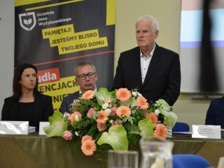 Siwowłosy mężczyzna przemawia za stołem z kwiatami na tle banera UJW.