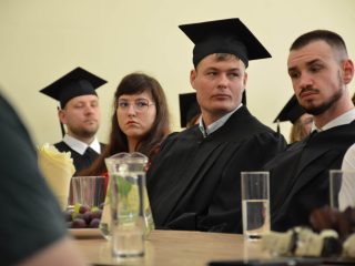 Absolwenci w czarnych togach i goście słuchają wystąpienia przy stole.