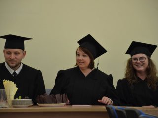Absolwent i dwie absolwentki w togach i biretach siedzą przy stole z poczęstunkiem.