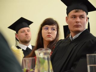 Dwaj absolwenci w biretach i kobieta w okularach spoglądają w stronę prowadzącego uroczystość.