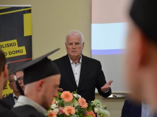 Siwowłosy mężczyzna gestykuluje podczas przemówienia przed absolwentami.