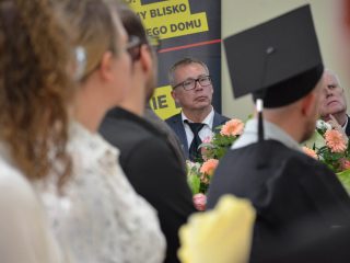 Mężczyzna w okularach siedzi za stołem z kwiatami, widoczny między absolwentami.