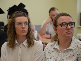 Dwie kobiety w okularach i jasnych bluzkach słuchają wystąpienia podczas wręczenia dyplomów.