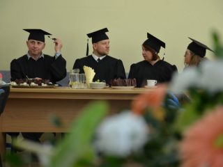 Czworo absolwentów w togach i biretach siedzi przy stole; jeden poprawia biret.