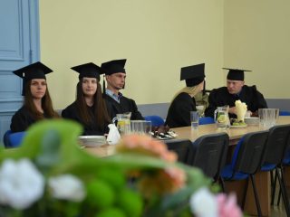 Absolwenci w togach siedzą przy długim stole, z kompozycją kwiatową na pierwszym planie.