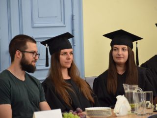 Dwie absolwentki w biretach siedzą przy stole obok brodatego mężczyzny w okularach.