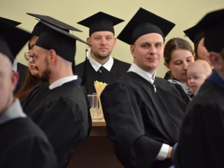 Absolwenci w czarnych togach siedzą wśród gości, w tym kobiety z małym dzieckiem.