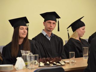 Dwie absolwentki i absolwent w togach siedzą przy stole z poczęstunkiem.