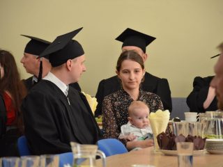 Kobieta trzyma niemowlę na kolanach obok absolwenta w todze i birecie.