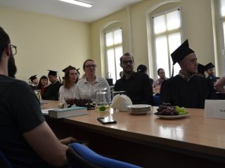 Goście uroczystości i absolwent w birecie siedzą przy stole z napojami i owocami.
