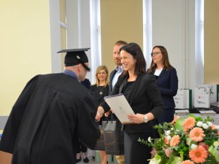 Absolwent w todze odbiera gratulacje od kobiety trzymającej teczkę z dyplomem.