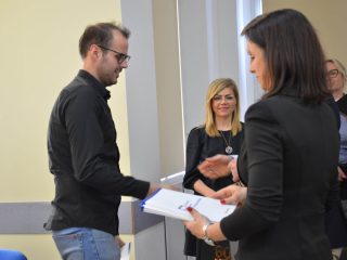 Mężczyzna w okularach i czarnej koszuli odbiera dokument od organizatorki uroczystości.