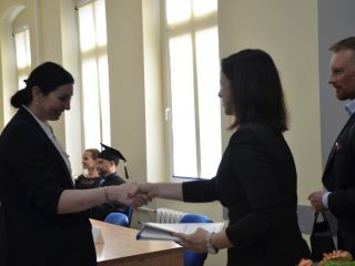Kobieta w granatowej marynarce ściska dłoń organizatorki trzymającej dokumenty.