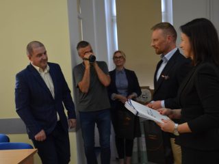 Uczestnik w niebieskim garniturze stoi przed organizatorami, a fotograf dokumentuje uroczystość.