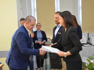 Mężczyzna w niebieskim garniturze w prążki odbiera gratulacje od organizatorki.