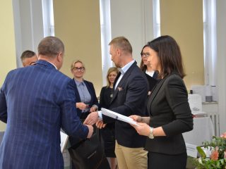 Mężczyzna w niebieskim garniturze ściska dłoń przedstawiciela wręczającego nagrody.