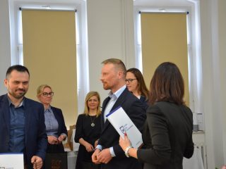 Mężczyzna z dokumentem odchodzi od organizatorów uroczystości wręczenia dyplomów.