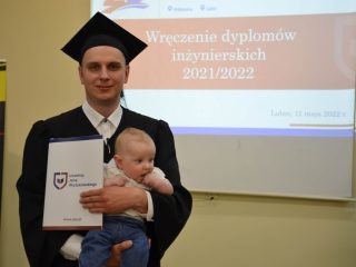 Absolwent w todze i birecie pozuje z teczką z dyplomem oraz niemowlęciem na ręku.