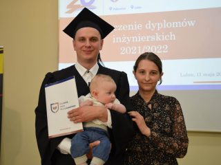Absolwent z dyplomem i niemowlęciem pozuje obok kobiety na tle ekranu uroczystości.
