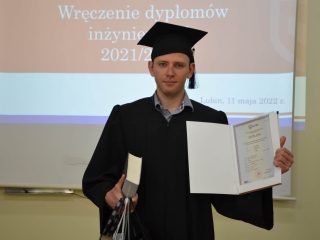Absolwent w todze prezentuje otwarty dyplom i unosi kciuk.